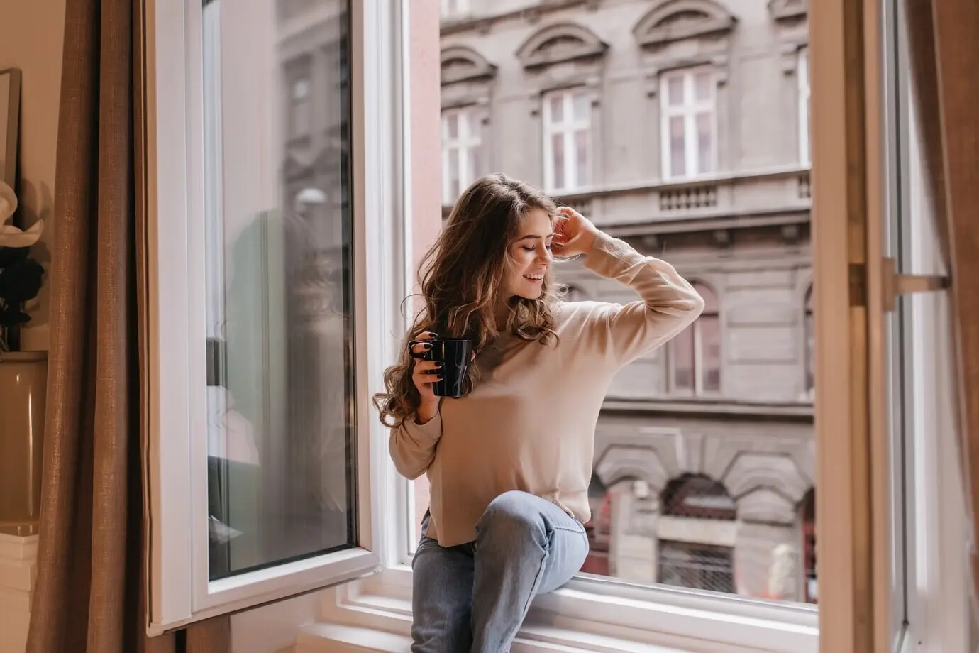 An elegant young woman in a beige shirt sits on a sill, looking at the city.