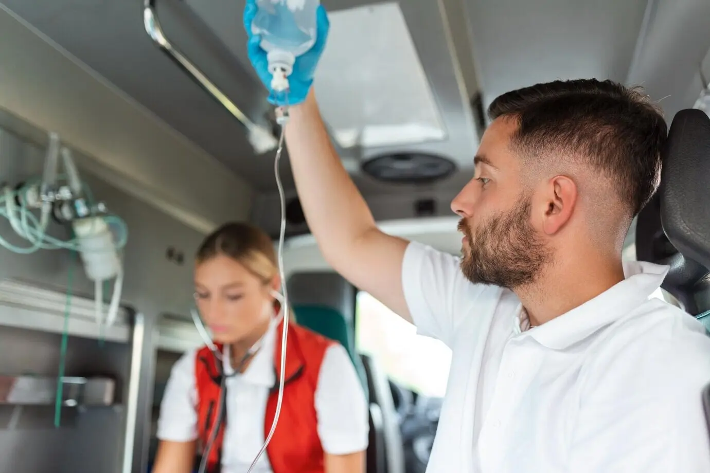 Medics attending an injured patient and administering an infusion; young male paramedics hold the IV solution, while a female doctor listens to the patient’s heart.